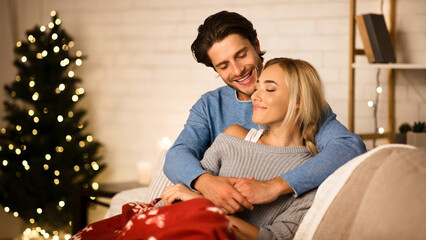 Cozy Christmas eve. Loving couple embracing near Christmas tree, resting on sofa