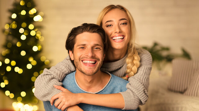 Happy couple looking at camera on new year's eve, woman embracing man in front of Christmas tree
