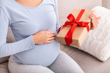 Unrecognizable Pregnant Lady Holding Wrapped Gift Box Sitting On Couch At Home. Cropped