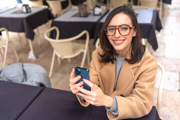 Happy middle-aged woman using smartphone sitting outdoor cafe. Smile businesswoman in glasses dressed trench coat holding smartphone in hand looking at camera. High angle view. Copy space