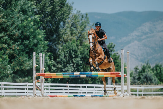 A focused rider and their horse jump over a colorful hurdle during an outdoor equestrian event on a sunny day.