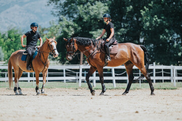 Obraz premium Two people riding horses in an outdoor equestrian arena, enjoying a sunny day. They are wearing helmets and riding gear.