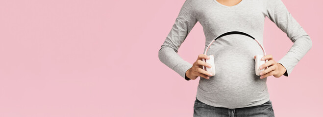 Pregnancy therapy. Unborn baby listening music in mother's belly. Womna holding headphones on her belly, pink background, crop