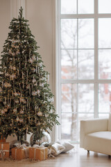 A large Christmas tree with many ornaments sits in front of a window. The room is decorated with a white couch and a few boxes. Scene is festive and cozy, with the tree and decorations creating a warm