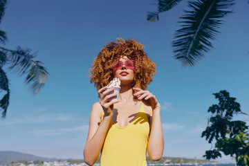 Young woman with curly hair enjoying ice cream against a clear blue sky and palm trees