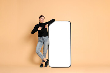 Beautiful young woman pointing at big mobile phone with blank screen on dark beige background