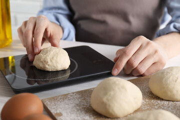 Woman weighing raw dough ball on kitchen scale at white wooden table, closeup