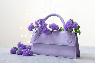 Stylish violet bag and ageratum flowers on gray table against light background