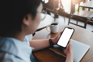 A woman is sitting in a cafe, holding a blank screen mock-up mobile phone in one hand and a coffee cup in the other...