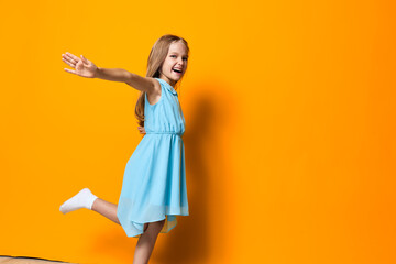 Joyful little girl in a blue dress leaps with arms outstretched against a vibrant orange wall...