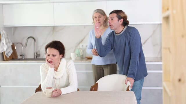 Old mother and man scold san woman who listens and sits with her back to them at table in kitchen 