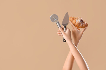 Female hands with spatula, pizza cutter and tasty croissant on beige background