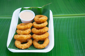 Golden onion rings served with sauce in biodegradable tray on green leaf.
