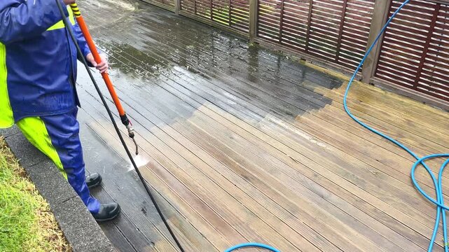 Man in waterproof overalls using a telescopic wand to water blast a wooden deck clean from built up grime and slime