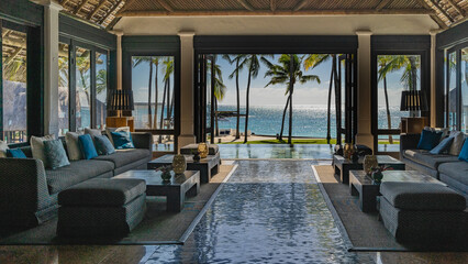 The lobby at the resort. Upholstered sofas with cushions, armchairs, tables stand along the glazed walls. Through the open doors you can see the turquoise ocean, the pool, the beach, coconut palms 