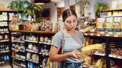 Smiling attractive young girl carefully comparing different types of pasta, deciding which to purchase while standing among grocery shelves in organic food store
