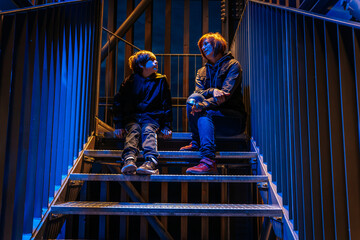 A mother and son sit together on metal stairs illuminated by blue lighting. They are engaged in a quiet conversation, surrounded by an industrial structure that adds a modern and intimate atmosphere.