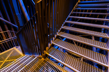 A detailed view of a metal staircase with grid-patterned steps, illuminated by blue and yellow...