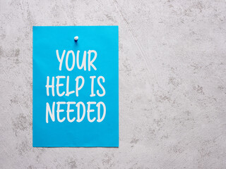 Your help is needed announcement on a blue sheet of paper pinned to a white concrete wall.