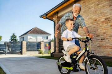 Happy grandson is learning how to ride a bicycle and his grandad is helping him.