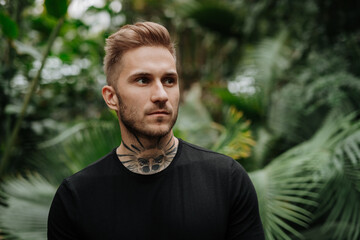 Handsome man with tattoo standing in botanical garden, in the middle of lush green foliage.