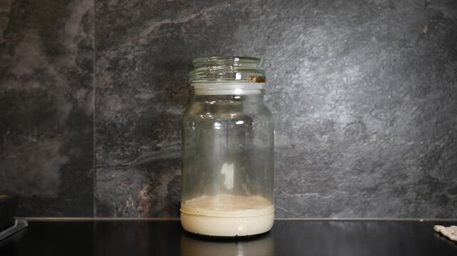 Timelapse of a sourdough starter during the fermentation process in a glass jar against a dark grey marbled kitchen counter and wall backdrop