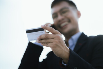 A businessman confidently using his mobile phone and credit card for online shopping or banking outdoors, embracing technology and e-commerce in a modern city setting.