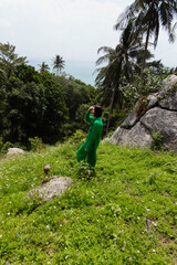 Fototapeta premium Person in green stands on grassy hill, gazing at lush forest, palm trees, rock, and ocean in the background