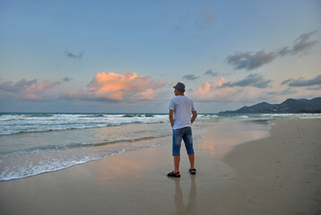 Man in white shirt, blue shorts, and hat watches sunset on sandy beach. Waves lap gently, sky is a mix of blues and pinks