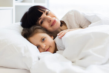Mother and daughter snuggling under white covers, sharing tender moment in bed. Warm, intimate scene displaying family love, comfort, and bonding in clean, cozy bedroom setting.