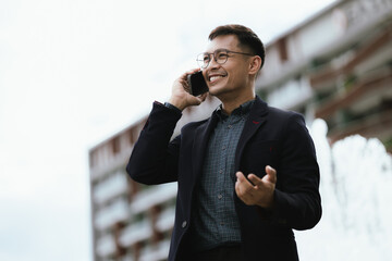 Confident Asian businessman in modern city, smiling while using smartphone outdoors, wearing professional suit, representing success and connectivity in digital age.