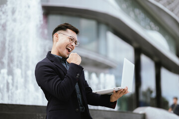 A determined young businessman in a city, confidently working on his laptop outdoors, dressed in a...