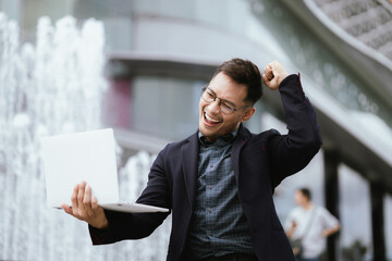 A determined young businessman in a city, confidently working on his laptop outdoors, dressed in a...