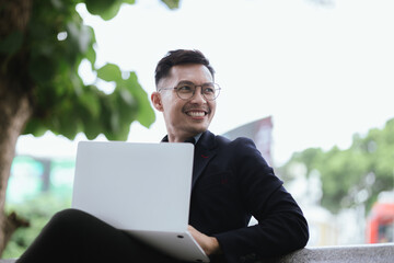 A determined young businessman in a city, confidently working on his laptop outdoors, dressed in a...