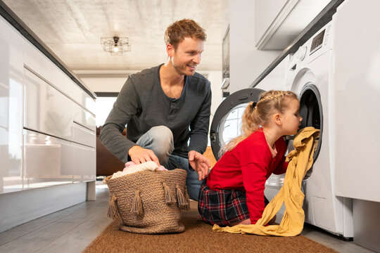 Father looking at daughter loading clothes in washing machine at home