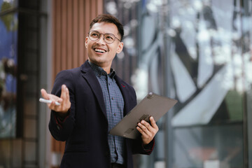 A cheerful young Asian businesswoman in a modern city, smiling confidently while using her smartphone outdoors, dressed in a professional suit, embodying success and digital connectivity.