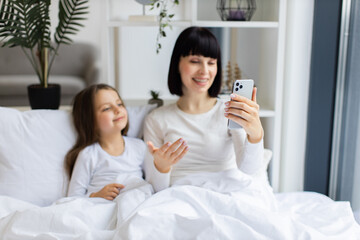 Mom and daughter lying in bed having video call talking on phone. They appear happy and relaxed, enjoying morning together.