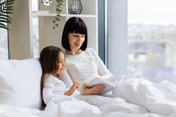 Mother and daughter enjoying quality time reading book in cozy bedroom. Bright natural light from window. Family bonding, love, and togetherness in comfortable setting.
