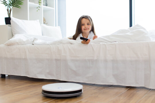 Smiling girl watching robot vacuum cleaner in modern bedroom with white decor. Concept of smart home technology, cleanliness, and eco-friendly living.