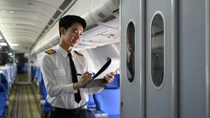 Smiling asian pilot standing inside an airplane, holding clipboard and reviewing or completing some paperwork