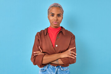 A woman with short silver hair stands with arms crossed, wearing a brown shirt and red top, exuding...