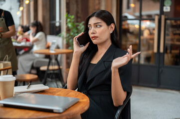 Obraz premium Uncertain, thoughtful Asian businesswoman is talking on the phone while sitting at a table of a cafe
