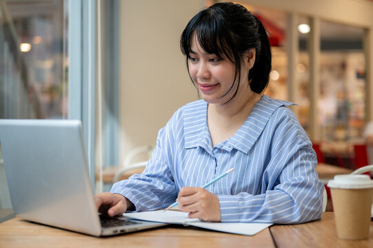 A young beautiful Asian woman is studying online via her laptop or working remotely from a cafe.