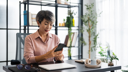 A middle-aged businesswoman with short gray hair is sitting at a modern desk, focused on her smartphone, with a calm and professional demeanor