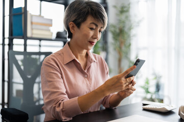 A middle-aged businesswoman with short gray hair is sitting at a modern desk, focused on her smartphone, with a calm and professional demeanor