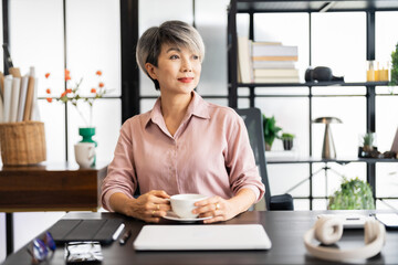 aged businesswoman with short gray hair is seated at her desk, looking thoughtfully upward while resting her chin on her hand in a modern, stylish office