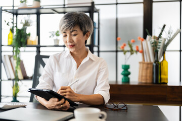 A middle-aged businesswoman is sitting at her desk in a modern office, working on a tablet with focus and concentration