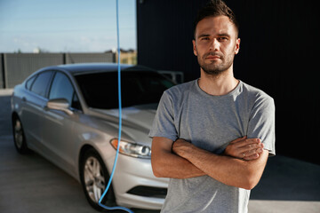 Arms crossed, standing. Man is with his car outdoors