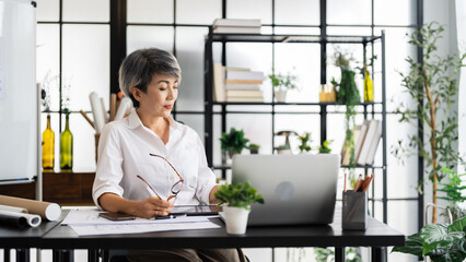 A female architect is deep in thought, holding a pen while reviewing blueprints at her desk, with...