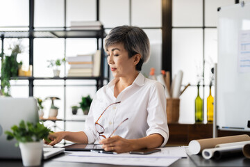 A female architect is deep in thought, holding a pen while reviewing blueprints at her desk, with...